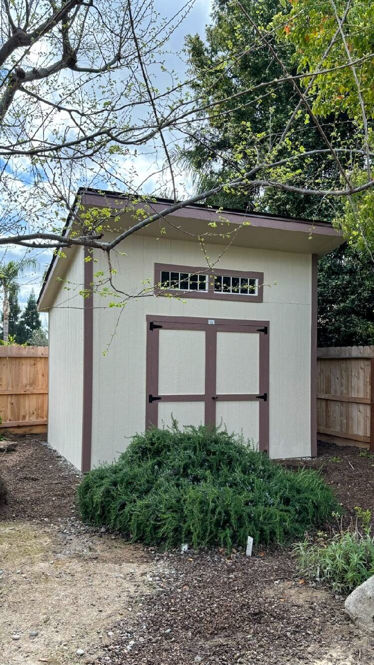 custom lean to sheds near Tulare CA with tan siding and brown trim and transom windows sitting in corner of fenced in area by bushes