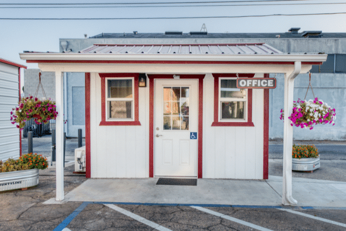 custom off white shed with red trim and metal roof featuring overhang office sign and hanging flowers