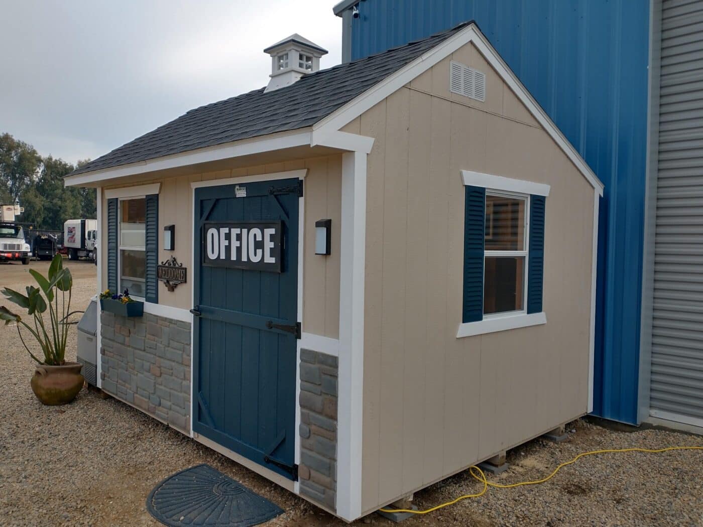 custom tan shed with office sign on blue front door gray detailing on bottom white trim and two windows