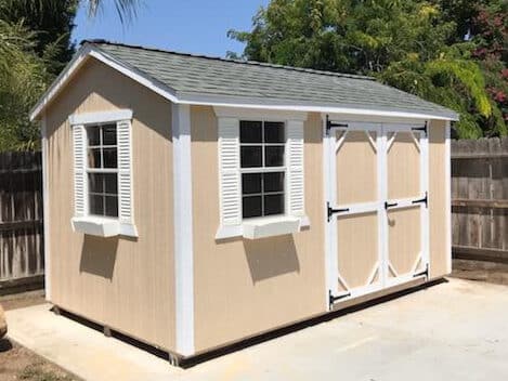 tan shed with white trim two windows and gray shingle roof on concrete pad by fence