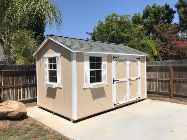 tan shed with white trim two windows and gray shingle roof on concrete pad by fence