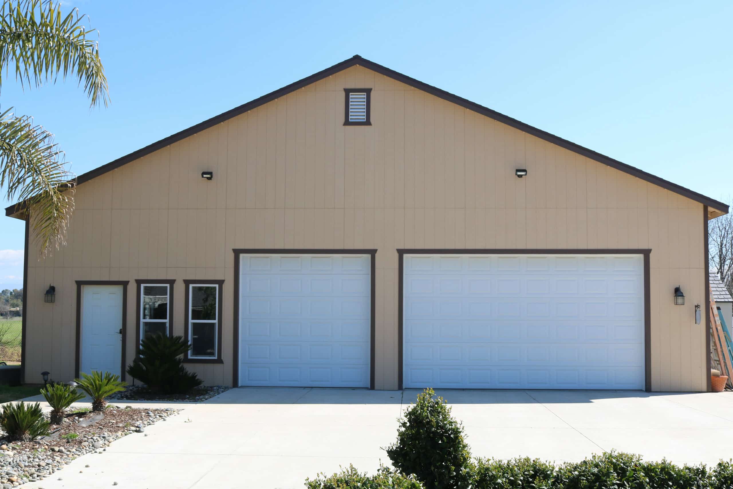 tan three car garage in Clovis CA with dark brown trim and side door surrounded by palm tree and other foliage