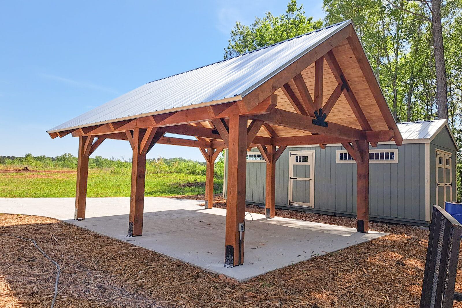 brown timber frame pavilion on concrete pad next to blue shed with white trim all on top of mulch