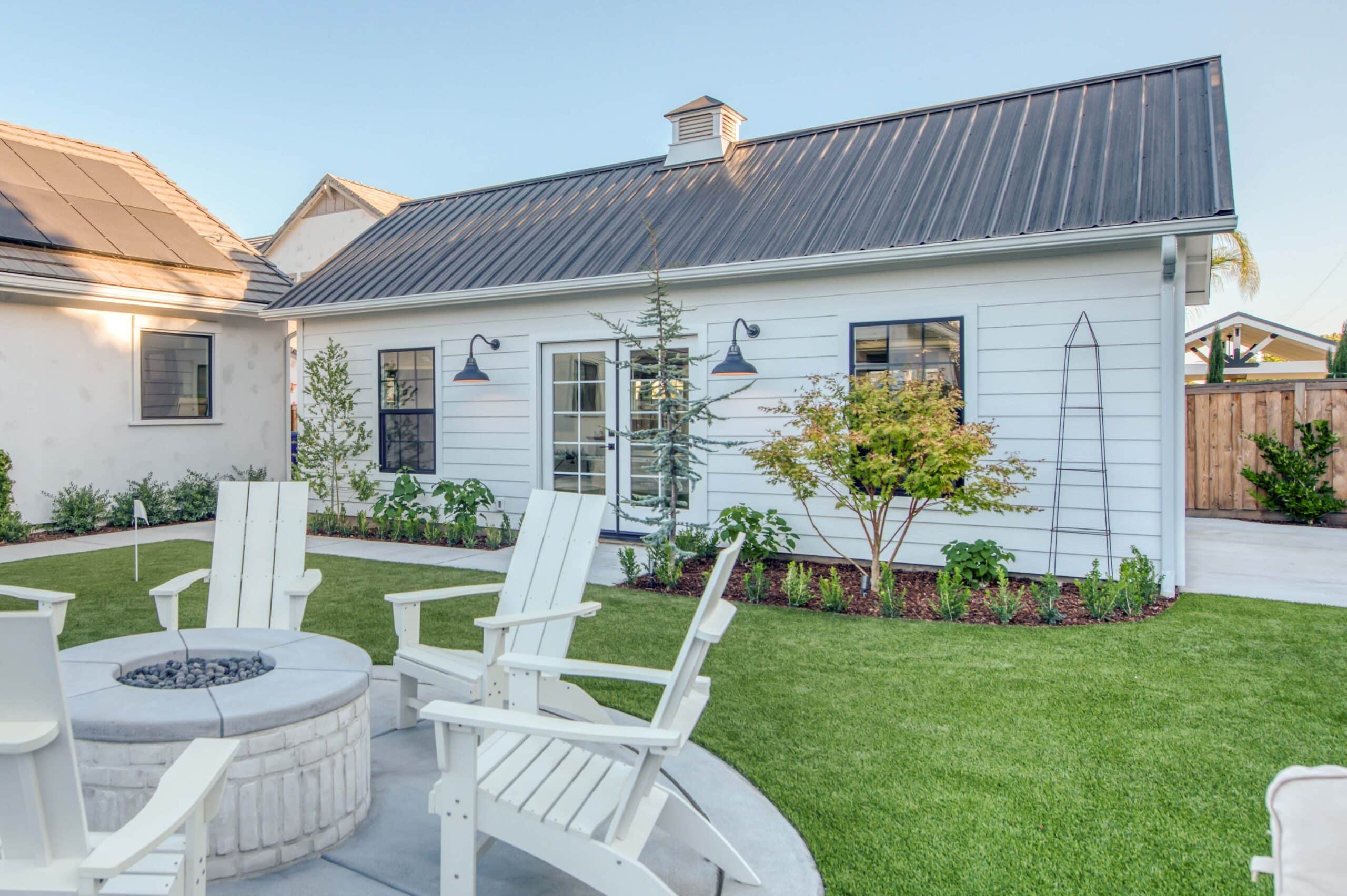 white cabana with french doors and garden in front by fire pit with 6 white chairs around it