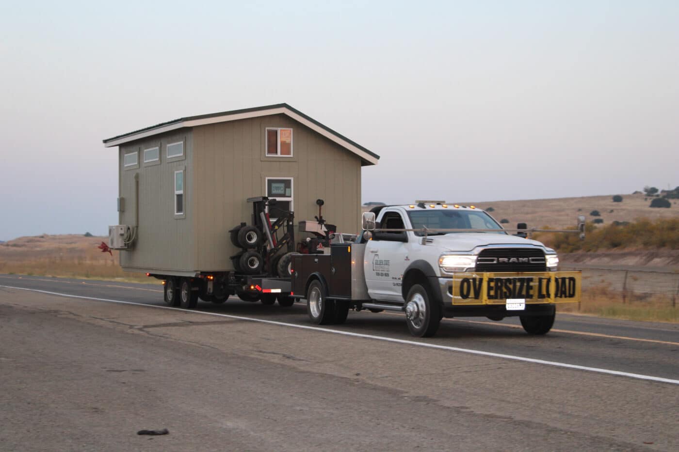 white truck with oversized load sign on front carrying tan shed down highway to deliver in Shaver Lake CA