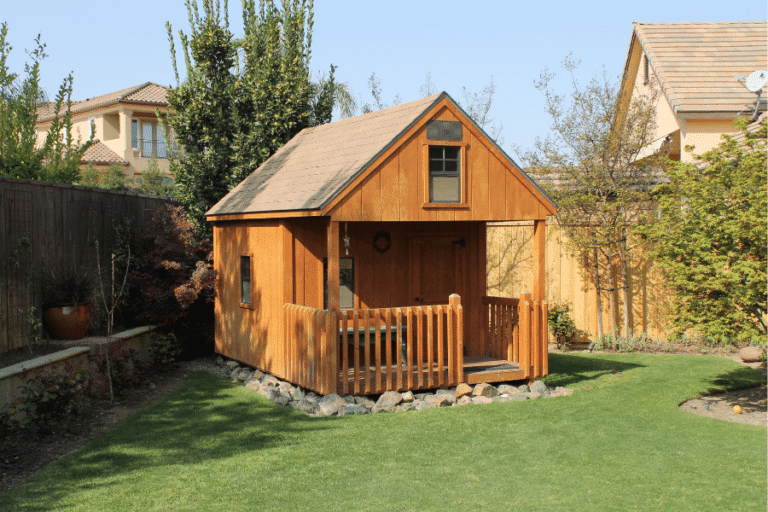 wooden playhouse with front porch on stones in corner of yard along fence and surrounded by trees