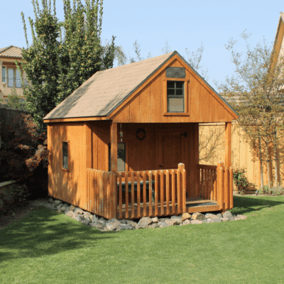 wooden playhouse with front porch on stones in corner of yard along fence and surrounded by trees